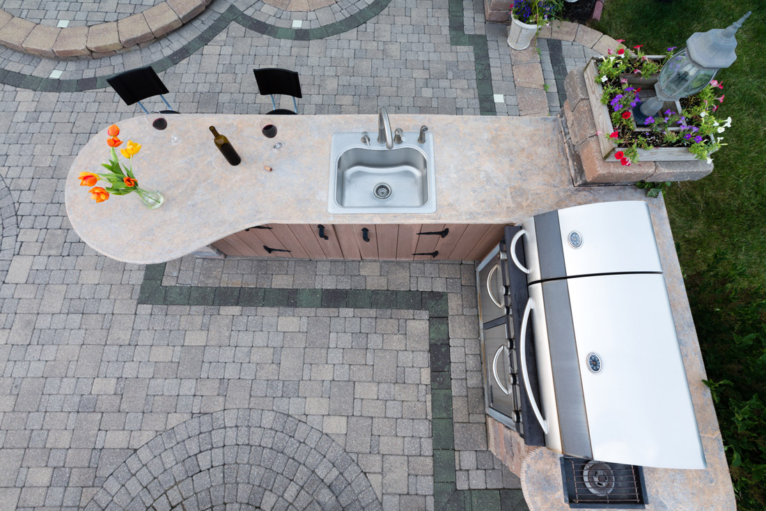 Outdoor summer kitchen with a built in gas barbecue and sink in a cement counter top in a corner of a brick paved patio for healthy living, view from above